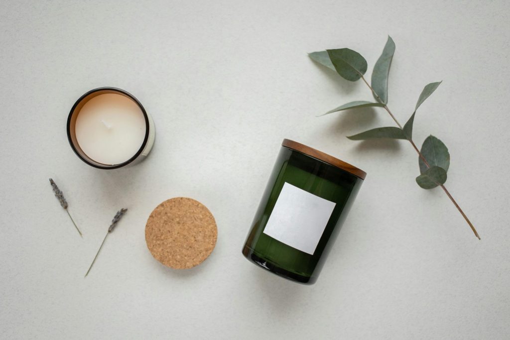 Top view of candles in green glass jars with leaves and cork lid on a white surface.
